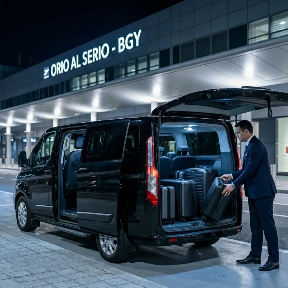 Un Van Ford Tourneo Custom nero di BasiglioTaxi con portellone posteriore e porta scorrevole aperti davanti al terminal di Orio al Serio di notte, con un autista che carica bagagli ordinati, in un'inquadratura quadrata Business Silver.